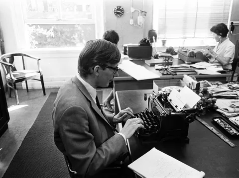 Fotografia d'epoca di un copywriter che batte a macchina in un ufficio pubblicitario, con una collega al lavoro sullo sfondo: il momento in cui una strategia diventa una frase scritta.