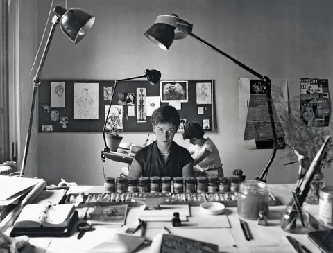 Fotografia d'epoca di un'illustratrice al tavolo da disegno, circondata da una fila di boccette di colore: la stessa tavolozza usata per registri diversi a seconda del momento.