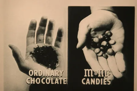 Un annuncio in bianco e nero con due mani aperte a confronto: una piena di scaglie di cioccolato comune, l'altra di confetti M&M's colorati, con le scritte «Ordinary chocolate» e «M&M's Candies»