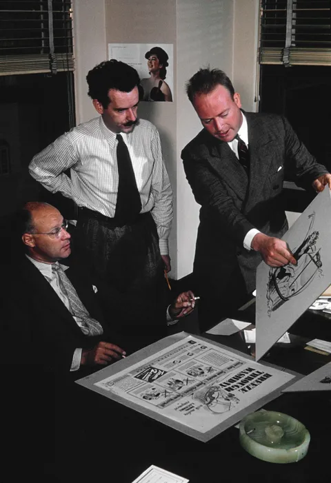 Fotografia d'epoca di tre persone in agenzia che discutono attorno a un tavolo, confrontando bozzetti e layout stampati di uno stesso annuncio pubblicitario.